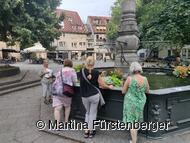 Das Bild zeigt den Brunnen auf dem Hafenmarkt. Um diesen stehen Personen und bei Ihnen steht eine Stadtführerin. 