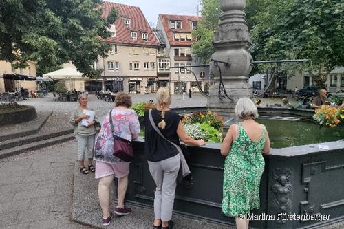 Das Bild zeigt den Brunnen auf dem Hafenmarkt. Um diesen stehen Personen und bei Ihnen steht eine Stadtführerin. 