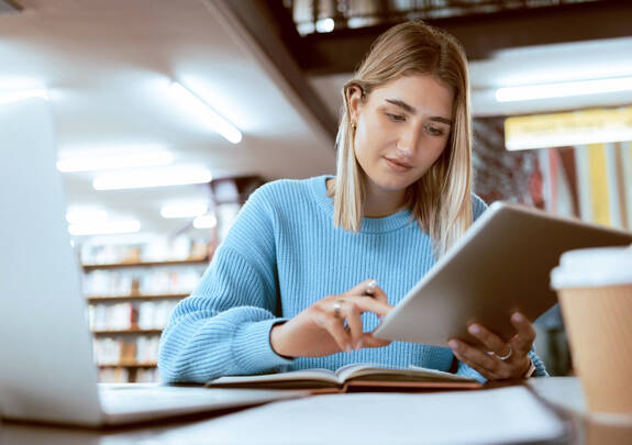 Eine junge Frau sitzt in einer Bibliothek und sieht sich etwas auf einem Tablet an